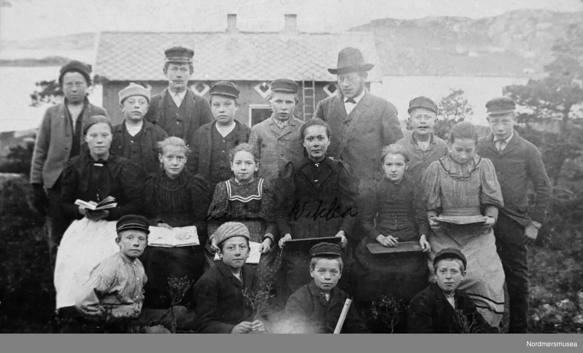 Gamle Bolgen skole før den ble flyttet til Valen i Vadsteinsvik. Nederst venstre:  Thomas Johansen, Harald G., Birger Schmedling, Søren Schmedling. Miderst venstre:  Signe Kristine Søberg, Lovise Schmedling, Nikoline Thorsteinsen, Ida Løkkemyr. Bakerst venstre:  Ludvig K. Bolgen, Alfred Sjølberg, Peder Nordhvalen, NN, NN, lærer Sivert Myhr, NN, Lars Sæter. Bolga tilhørte på denne tiden dette bildet ble tatt, Bremsnes skolekrets og kommune.  Bilde fra fotoinnsamlinga i Frei kommune 1978-79. Innsamla og registrert av Freielever på Nordlandet ungdommskole. Fra Nordmøre Museum sin fotosamling. Reg: EFR
