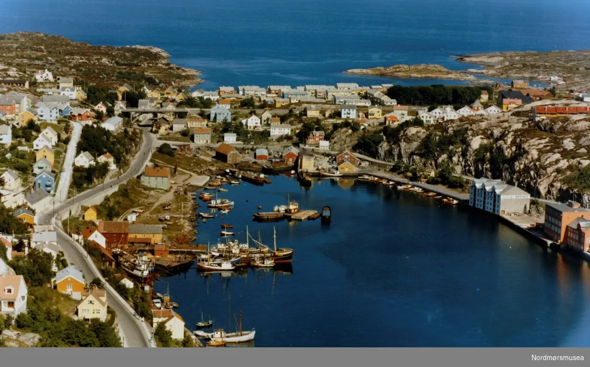 Flyfoto fra Vågen i Kristiansund, med Kirkelandet på venstre side og Gomalandet på høyre. Her ser vi blandt annet Mellemværftet med flere fartøy fortøyd like ved på Kirkelandet side, som i dag er en del av Nordmøre museum. Flyfotoet er utført av Widerøes flyveselskap den 17. juli 1965. Fra Nordmøre museums fotosamlinger. Reg: EFR
