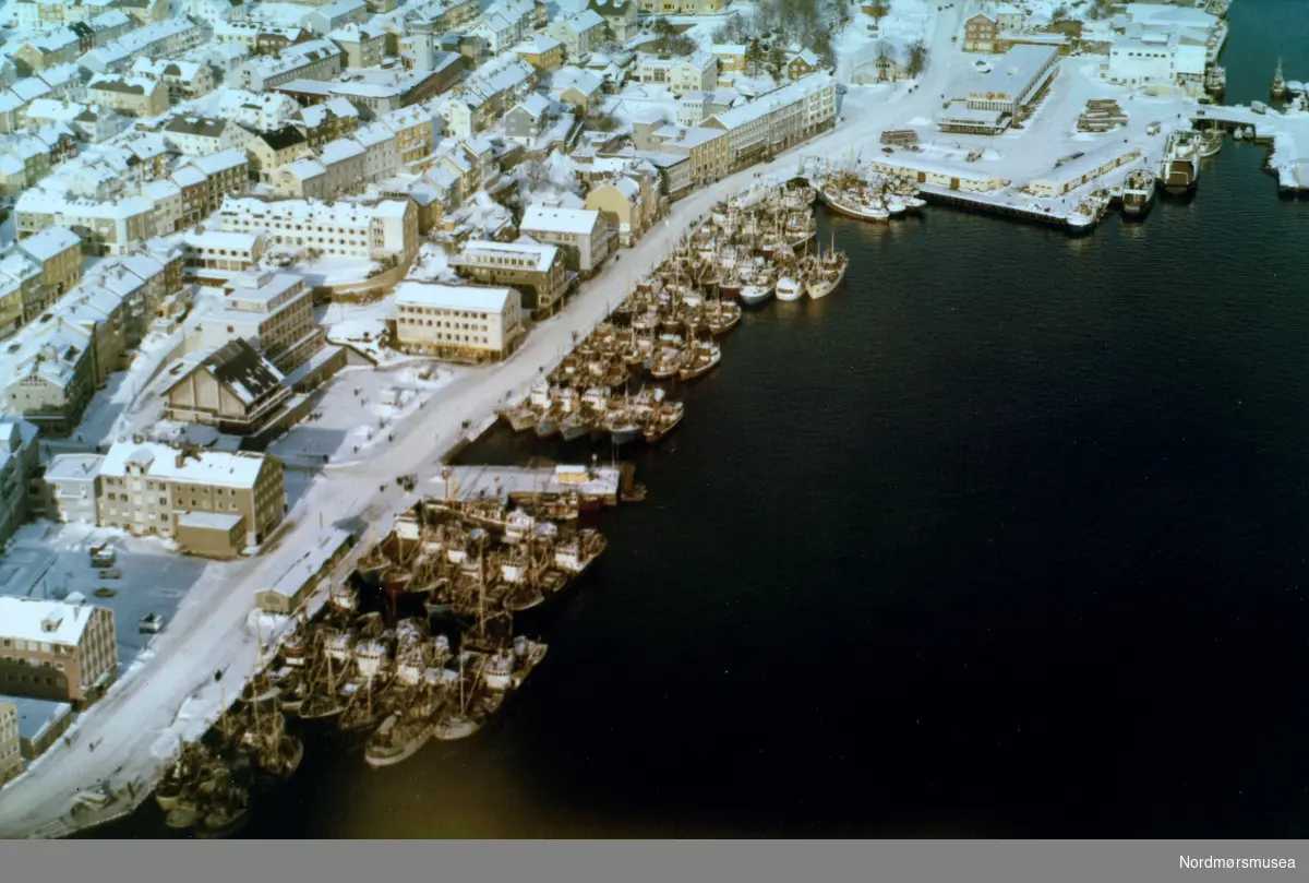 Flyfoto fra et snøkledt Kristiansund, hvorfra vi ser Rådhuset og Piren, Vågekaia med Vågeveien og Nordmørskaia og litt av Devoldholmen i bakgrunnen. Flyfotoet er utført av Widerøes flyveselskap den 23. februar 1969. Fra Nordmøre museums fotosamlinger. Reg: EFR
