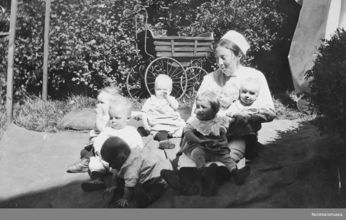 "Tage, Sissy, Imaatten, Kissen Jenny, Aslaug og Petter Kaare 1916" Foto av en barnepleier, her sammen med en gruppe barn. Trolig ved et barnehjem i Bergen. Teksten til bildet er meget utydelig og usikker. Albumet bildet er hentet fra har tilhørt Marit Hestvik, og viser bilder fra familien Rødsand/Hestviks fotoalbum fra Kristiansund. Det ble gitt til Nordmøre museum av Nelly Rødsand 5. september 1983. Fotoalbumet består av bilder med reg. nr. KMb-1983-024.0049 til KMb-1983-024.0223. Flere bilder fra andre album er å finne på samlingsnummer KMb-1983-024. Fra Nordmøre museums fotosamlinger. EFR2015