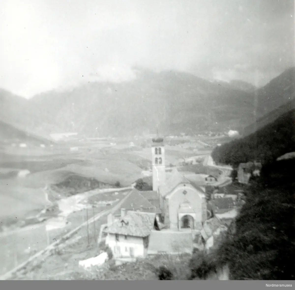 Foto fra ei bygd omkranset av fjell en ukjent plass. Er dette i Norge?
Fotograf er sannsynligvis noen fra Fasting familien. Fotoserie. Fotografier gitt til Nordmøre Museum fra Kate og Kristian Fastings arvinger per 1986. Fra Nordmøre Museums fotosamlinger.