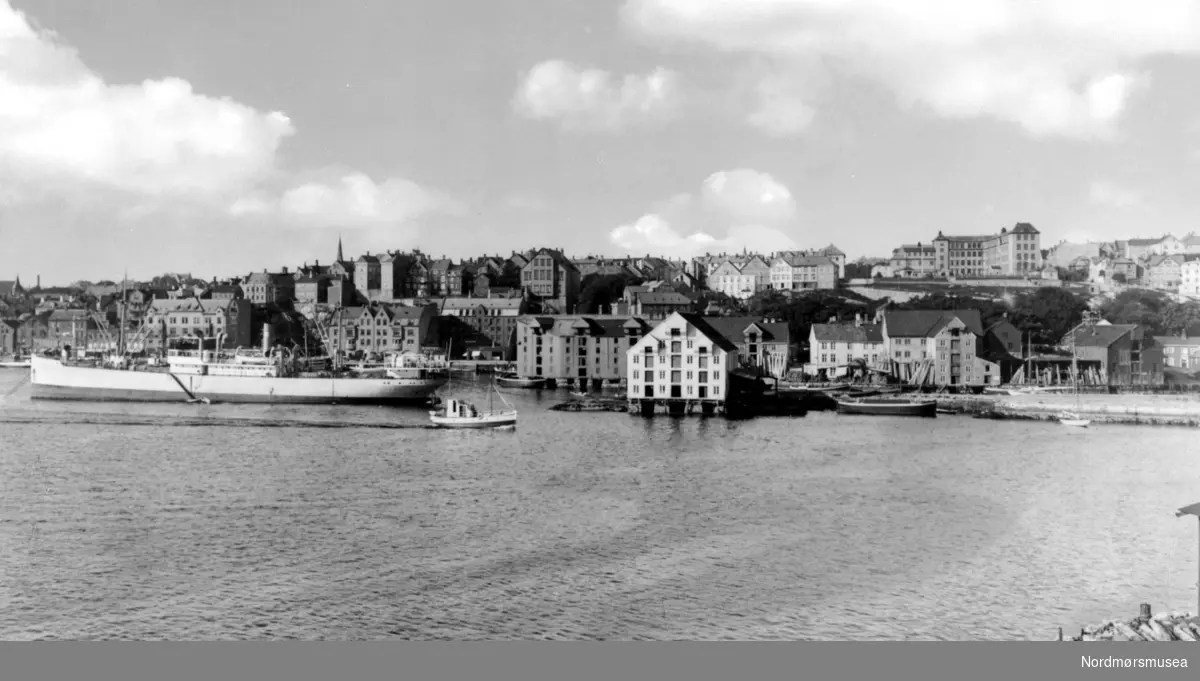 Foto fra et frakteskip nær Vågekaia/Johnsenhuken/Nordmørskaia, med Kirkelandet i bakgrunnen. Legg ellers merke til Allanengen skole i bakgrunnen på høyre side - som ble bygd i 1919. Postkortet/fotoet er forøvrig utgitt av Georg Sverdrup, og er fra 1936. Fra Nordmøre museums fotosamlinger. Tilleggsinfo: Bildet er tatt fra Israelsneset på Gomalandet. Lasteskipet ligger i ";storbøya"; ut fra Knudtzonbrygga (den hvite midt i bildet) på Knudtzonholmen, senere ";nye Devoldholmen";. Til venstre for den ser vi den store Astrupbrygga. Til høyre ligger Klingenbergeiendommen. Helt ute til høyre skimter vi den eldste av våningshusene på Fosna gård, mens det hvite mønet på den nye skimtes over brygga. - Over Klingenberg ser vi Allanengen skole. - Utfyllingen mellom holmene (Knudtzon og Devold) øst for Holmasundet er også godt i gang. (Informant: Tor Arne Skålvik.)



