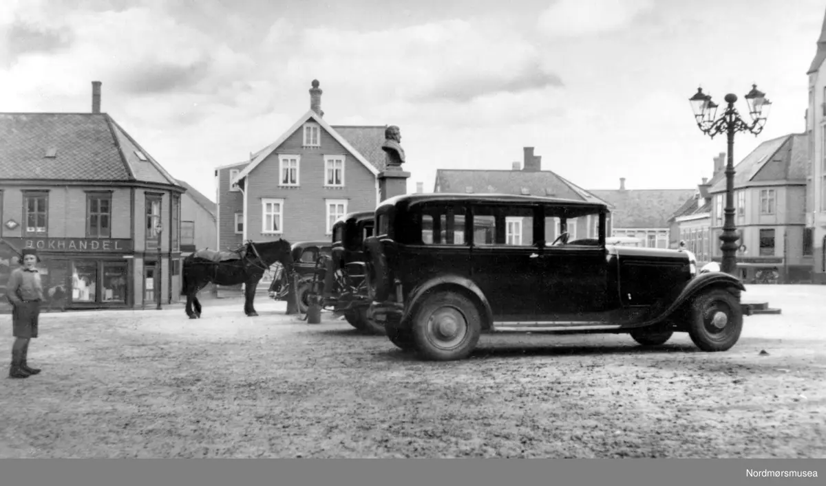 Til dateringen: Bakerste bil er mer avrundet, typisk 1933-34. (Info:
Ivar Stav) -  På bildet ser vi en rekke med biler stilt opp på det gamle Torvet i Kristiansund, foran Christiebysten som vi ser midt på bildet. her er hotell, bokhandel, kjøretøy, lys, byste, hest, dyr etc. Til høyre ser vi ";lysekrona";, navnet som den var kjent under i byen. Det var en flott støpejerns gatelykt som var smidd ved en av byens skipsverft. I moderne tid gjenreist foran Festiviteten og brukt som logo for Operaen i Kristiansund. I bakgrunnen ellers ser vi Sverdrups bokhandel til venstre, klesbutikk og Frisør i midten, mens til høyre ser vi litt av det gamle Grand Hotell (nr. 2) som var en flott murbygning, oppført i Jugenstil. Fra Nordmøre Museum si fotosamling.