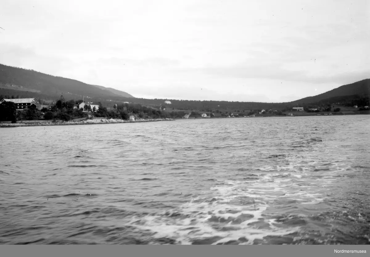 Tingvollvågen. Fjøs/låvebygning og våningshus i prestgarden til venstre. Kyrkja ligg så vidt utafor venstre bildekant. Kan det vera Vonheim sanatorium vi ser oppi skogen nær midten av bildet? (Info: Lars Brubæk).  Møre og Romsdal. Fra Nordmøre museums fotosamlinger.