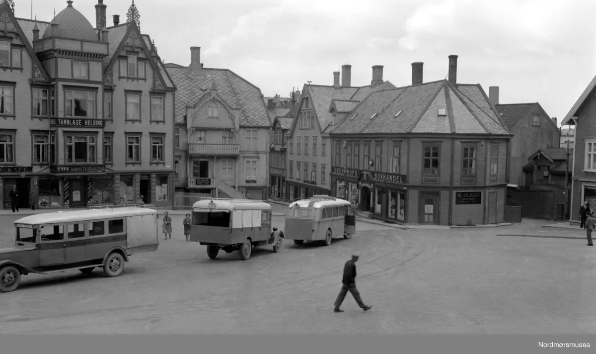 Foto fra torget på det gamle Kirkelandet, hvor vi blant annet ser tre busser . Vi ser også gården til tannlege Helsing i bakgrunnen, samt  Sverdrups bokhandel i bakgrunnen. Ellers legg merke til mannen som går over torgplassen i front. På bilen til venstre kan vi skimte Kristiansund-Oppdal Auto (KOA) på stripa under vinduene. Her er Oppdal stavet med P. Dette er av de tidlige rutebilene fra KOA. I 1932 åpnet ferjesambandet Kristiansund-Kvisvik. KOA og Surnadal Billag sine rutebiler kunne dermed følge ferja til Kristiansund sentrum. Det var først i 1938 at rutebilene fra Molde og Gjemnes kunne følge ferja til Kristiansund. Det var dette året ferjesambandet til Gjemnes og Torvikbukt åpnet. Bildet er derfor tidlidst fra 1932. (Info: Sveinung Berild - 11.11.2016). Fra Nordmøre museums fotosamlinger.