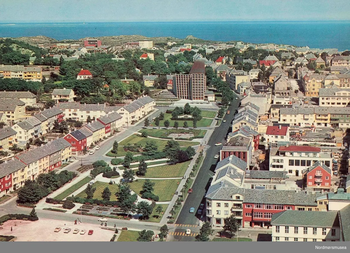 Postkort fra Parken på Kirkelandet i Kristiansund, hvor vi ser Kirkelandet kirke ligge i enden, og i den delen som heter Roligheten. Kirkelandet kirke ble innviet 24. mai 1964. Ellers så ser vi Langveien som går loddrett til høyre for parken, mens det er Konsul Johnsens gate som går på venstre side. Tverrgaten i front er Kong Olav V gate (per 1992) på venstre side av Langveien, og Skolegaten på høyre side. Kortet er utgitt av Widerøe's Flyveselskap A.S, Oslo. Fra Nordmøre Museums fotosamlinger.
