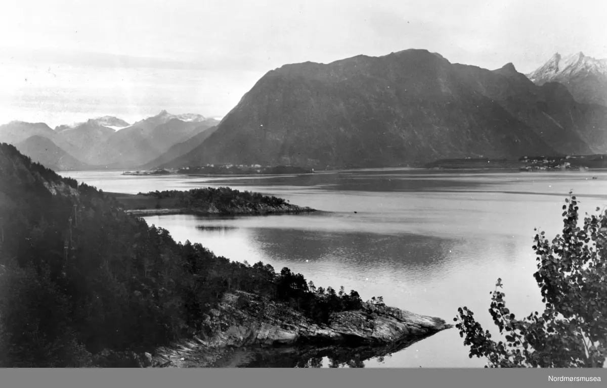 Dette bildet er tatt fra Børga i Torvik i Rauma kommune. Innover mot Åndalsnes til venstre, og Veblungsnes til høyre. Fjella til høyre er Vengetindene, og den mørkere Blånebba. Lengst inne i Isfjorden ser vi Juratind mot himmelen. Fjellet Aksla, eller Nesaksla, ovenfor Åndalsnes, midt imot. (Info: 
Eyolf Sæbø) - Landskapsbilde, hvor vi ser store fjell rundt en fjord. Ved foten av fjellet midt på bildet skimtes en bygd.  Møre og Romsdal omkring 1920 til 1939. Fotograf er trolig Georg Sverdrup. Fra Nordmøre museums fotosamlinger.
