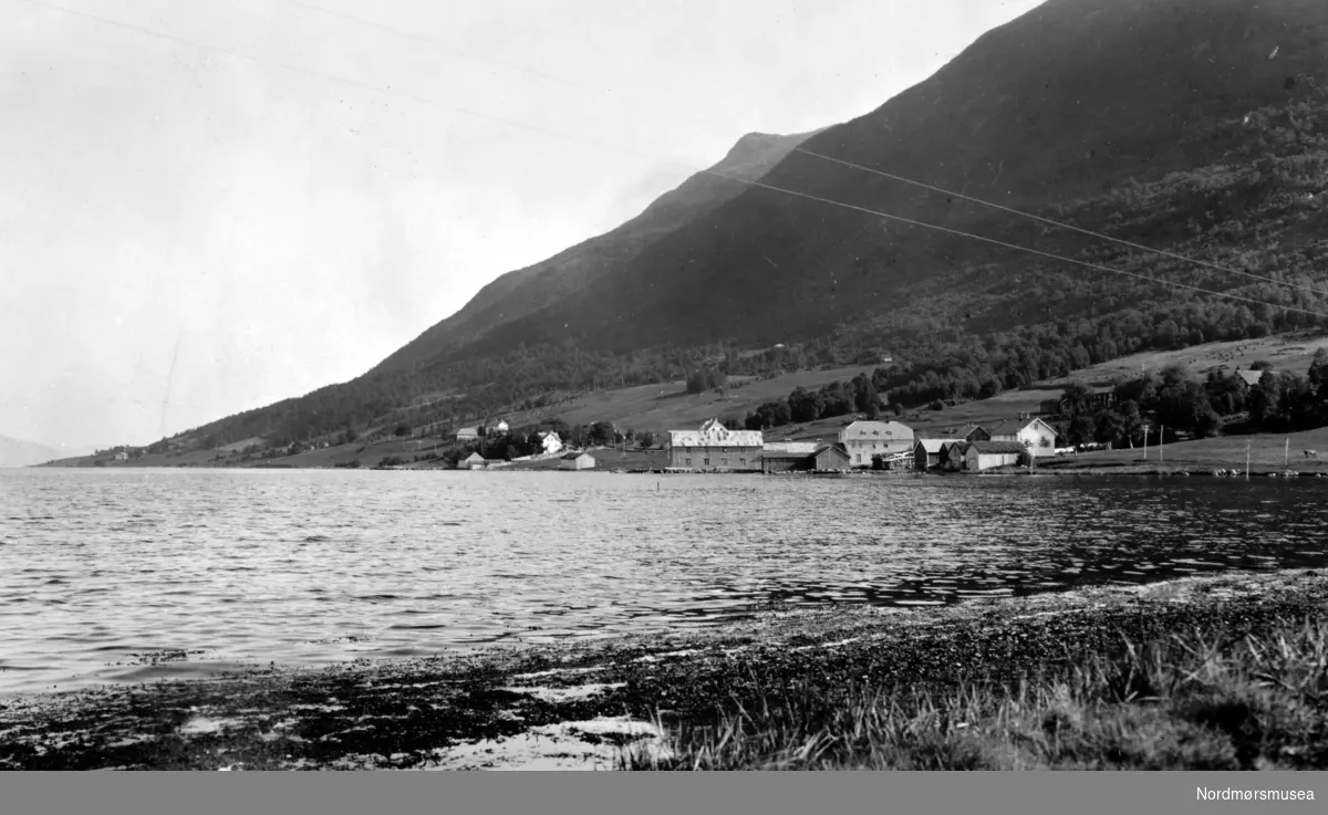 Foto med parti fra bygda Batnfjordsøra i Gjemnes kommune. Vi ser deler av bebyggelsen nede ved strandlinjen. Like bak ser vi de store fjellene som ruver godt i landskapet. Fotograf er Georg Sverdrup, og datering er 1935. Fra Nordmøre Museums fotosamlinger. Reg: EFR
