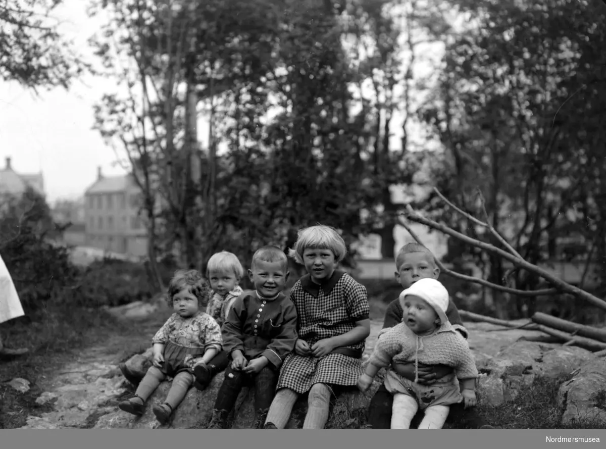 Foto av seks barn sittende ute på en liten fjellhammer, med trolig en barnevakt stående like ved til venstre. Dette er barnebarn av Margrethe og Georg Sverdrup, på bildet kan vi bl.a. se Liv og Kirsten Sverdrup , døtre av Nanna og Helge Sverdrup, og Finn Resvoll, sønn av Hildur Sverdrup Resvoll.
Bildet er trolig fra området ved Roligheten og familiens bolig i Rosentræders gate 1 på Kirkelandet i Kristiansund. Datering er sannsynligvis omkring 1928-1930. Fra Nordmøre museums fotosamlinger.
