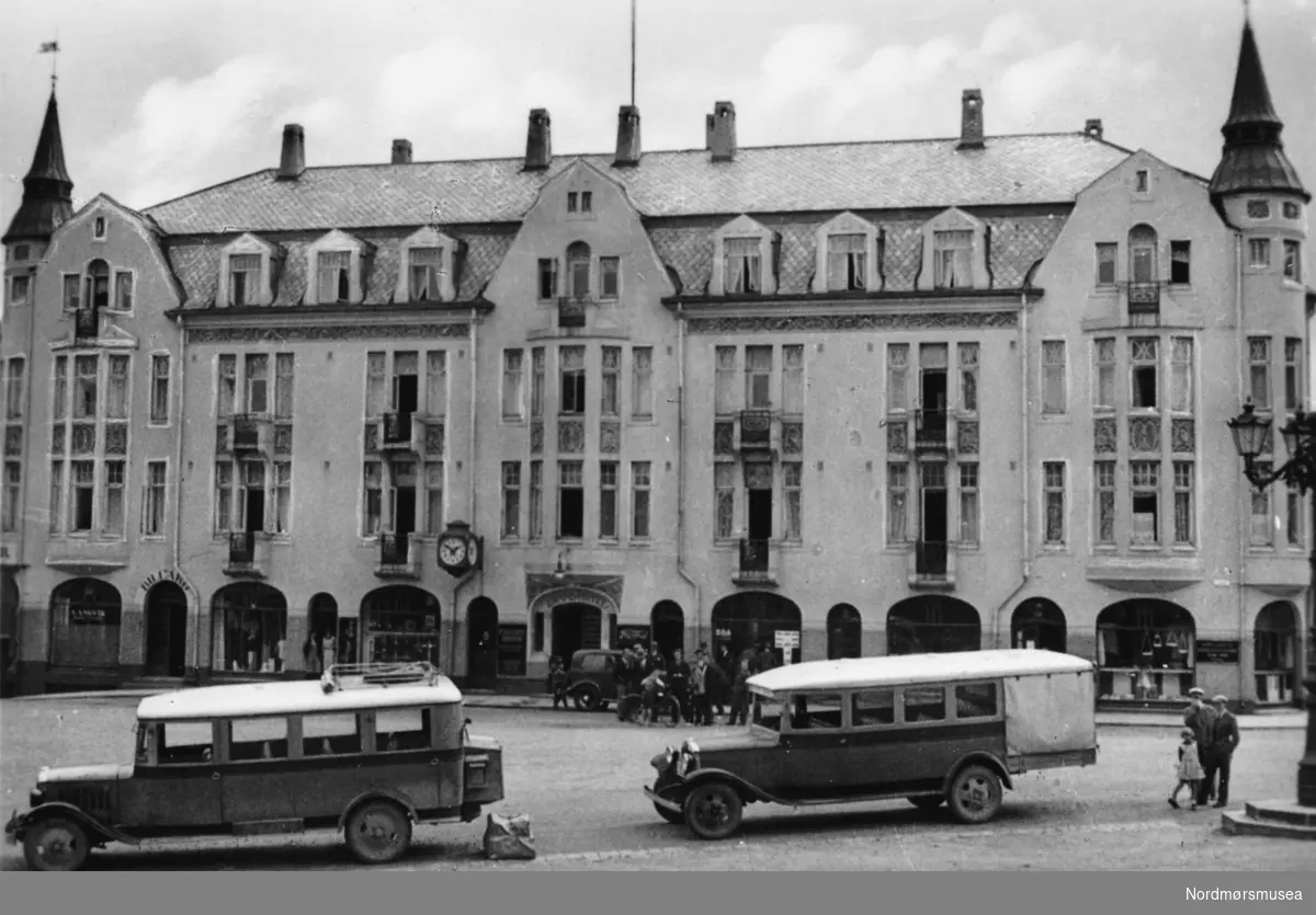 På bildet ser vi byens Grand Hotell nummer 2. Byens første Grand Hotell var bygd i Sveitserstil, men 8. november 1907 brant dette ned til grunnen. Eieren av Hotellet Nils Nilsen valgte senere arkitekt Tobias Rivertz fra Kristiania til å bygge opp hotellet. Rivertz oppholdte seg da i perioden 1907-1909 i Kristiansund, hvor han blant annet har tegnet ";en Badeanstalt";, Festiviteten, Grand Hotell og Norges banks lokaler i samme gaten som Grand Hotell. Hotellet ble oppført av murmesterfirmaet Olsen &amp; Michalsen i Jugendstil, og under  Rivertz' ledelse og tegninger. Hotellet kostet litt over 200.000 kroner og bygge, men summen inkluderer ikke tomten og monteringen (Referanse: En oplevelsens festlabyrint av Egil Husby, side 15 - 17). Nordmøre Museums fotosamling. Reg: EFR
