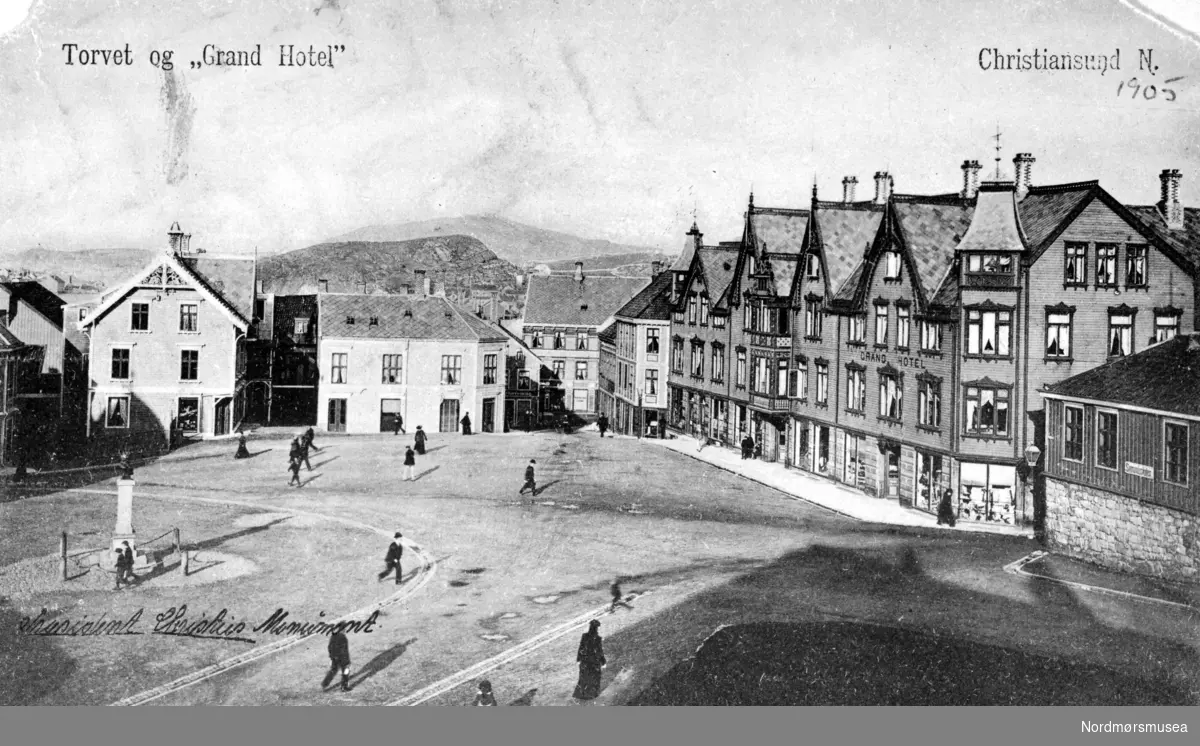 Foto fra torget på Kirkelandet, hvor vi ser byens Grand Hotell, bygd i sveitserstil, til høyre. Ellers så ser vi litt av Bernstorffskolen til høyre for hotellet. Skolen ble vedtatt revet i 1905 for å gjøre plass for Norges Bank, som vil bli reist på denne tomten i 1909 - samme året som Grand Hotell ble gjenreist etter en brann i 1907. i Christiebysten sees sentralt plassert på torvet til venstre. Denne er per 2008 å finne i President Christies gate, ved Blixhammeren. Motivet/postkortet er datert etter påskrift 1905. Fra Nordmøre Museums fotosamlinger. Reg: EFR
