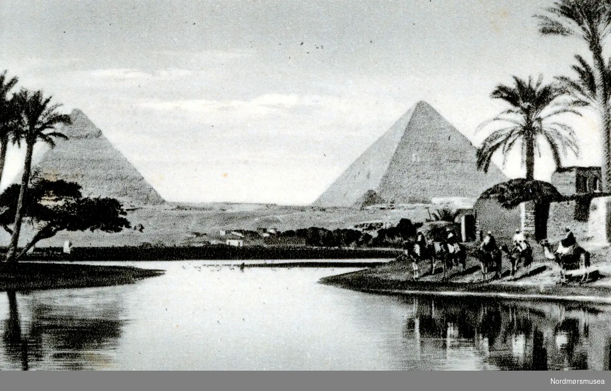 Foto av en gruppe nomader på kameler ved en innsjø i Egypt, med to pyramider like i bakgrunnen. Dato, sted og fotograf er ukjent. Fra Nordmøre museums fotosamlinger.
