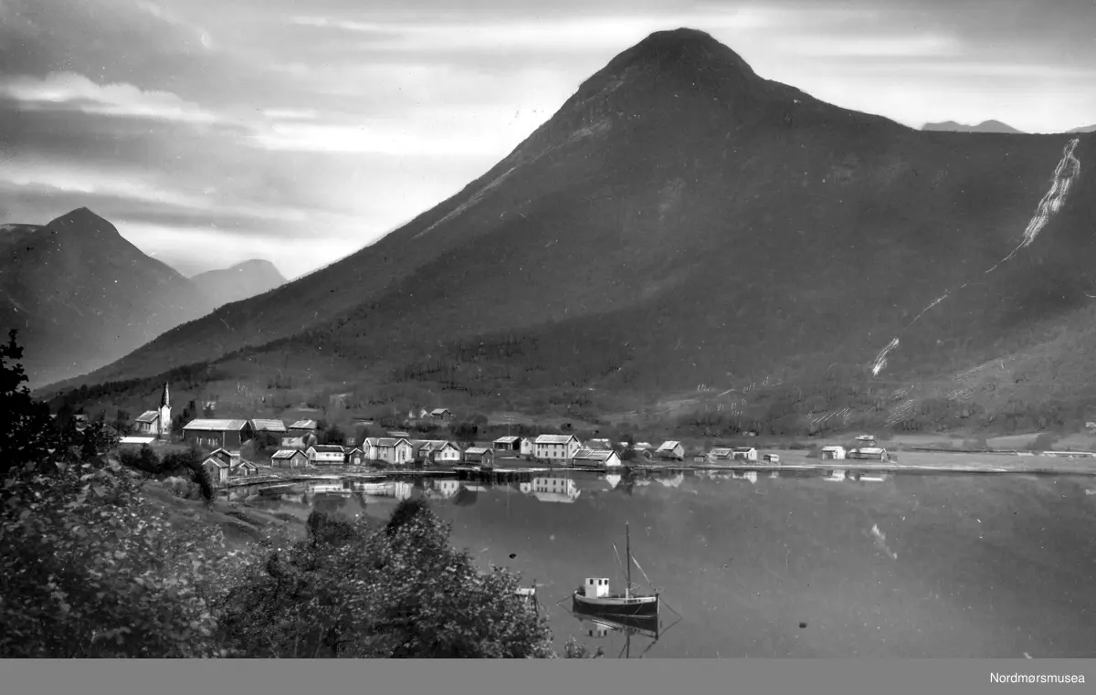 Dette er fra Øksendal i Sunndal kommune på Nordmøre. Vi kjenner igjen Storhaugen og fossen i Erstadelva. Likedan den spesielle, tette bebyggelsen ved sjøen. Øksendal kirke er fra 1894, og restaurert i 1972. Fjellet bak til venstre må være Kleppen. (E Sæbø) - 
Øksendalsøra. Fra Nordmøre Museums fotosamlinger.
