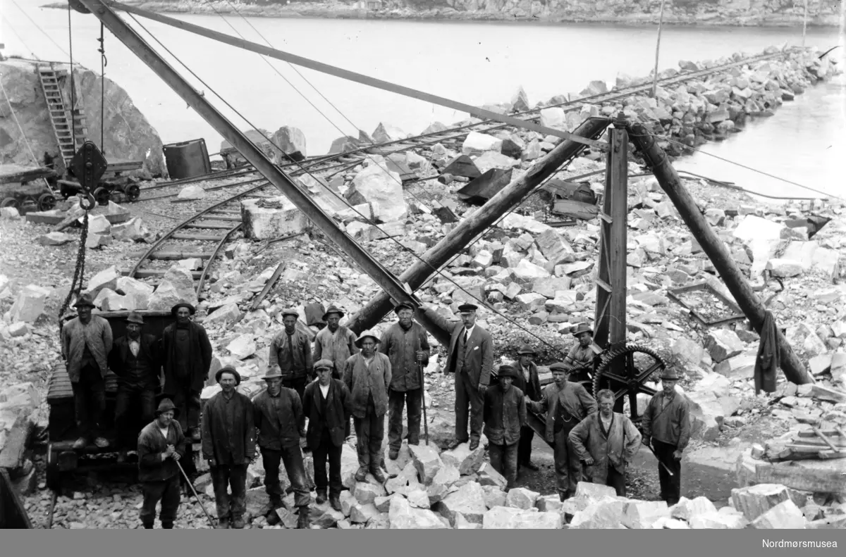 Anleggsgjengen samlet ved kranen på Innlandet under byggingen av moloen i Sørsundet. Ellers så ser vi tre menn stående på muligens en dressin på jernbanesporet som går til enden av moloen. Kristiansunds historie nevner at det helt fra havnefogd Schjetnans dager, 1890-årene, hadde det vært et ønske om en molo i Sørsundet for å forbedre forholdene. Behovet for dette ble virkelig satt i fokus da saltskipet ";Norden"; fikk føle hvor vanskelig forholdene kunne være da det grunnstøtte i Lervika vinteren 1903. På foto KMb-2008-026.0045 kan en se noe som ligner på en fylling, og som sikkert er den samme som en kan se på KMb-1941-000.0200, men vet ikke noe om det er denne fyllingen som er forløperen til Sørsundmoloen. Den ble vel bygget på 1930-tallet. Dersom det finnes stoff fra Havnekommisjonens arkiv kan det antakelig være noe å finne der. Fra Nordmøre Museums fotosamlinger.

ALTØ: Ut i fra Kristiansunds historie så kommenterte Romsdalsposten i 1917 moloen på Innlandet med at en måtte ha i tankene at en måtte både ha framtidig bru og ";jernbanespor fra Indlandet til stationen i Øvervaagen";: ";Der maa takes større maal og sees længere frem, naar man nu skal foreta reguleringer og utbygninger";.

Ser ut for at moloen enda ikke var påbegynt, men i Kristiansund historie står det at

";Siste ledd var (utbyggingen) Innlandets tilknytning til detnye vannverk i 1920.";

Moloen var ikke med i dette arbeidet.

I tiden rundt 1922 i forbindelse med nødslån blir det kommentert at

";De mest synlige resultater av nødsarbeidet var ellers de betydelige utvidelser av kaiene - eller ";almenningene";, som de ennå ble kalt - på Innlandet, og enda mer på Nordlandet. Planer om et moloanlegg i Sørsundet, utført for Havnedirektoratets regning, hadde vært fremme helt siden ";Nykaia"; på Kirkelaneet sto ferdig under verdenskrigen. Arbeidsledigheten gjorde et slikt prosjekt særlig aktuelt, og det lyktes så smått å komme i gang. Men bevilgningene stoppet opp etter et par år, og etterlot bare ";en veldig stenrøis";.

Reknergata og Heinsagata ble opparbeidet i denne perioden.

Moloen ble altså ikke ferdig da heller.

Fotoene som er i museets samling er mye sannsynlig fra 1920-tallet.



