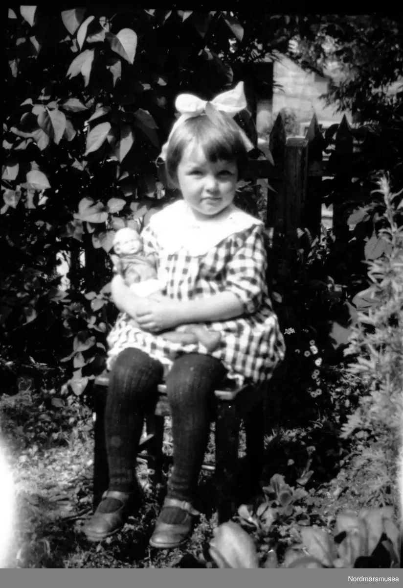 Foto av en liten pike sittende ute i en hage sammen med dukken sin. Barnet er trolig fra Rødfamilien, og det samme er trolig fotografen. Ukjent datering, trolig mellom 1930 til 1939. Giver av bildet er Turid Rød. Fra Nordmøre Museums fotosamlinger. 
Reg: EFR



