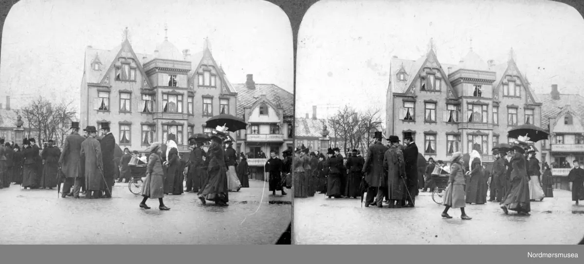 Stereobilde fra Torget av Kristiansund, hvor vi ser en større folkemengde samlet på torgplassen en regnværsdag. De fleste med penere klær, så bildet er trolig fra en søndag eller en hellidag. I bakgrunnen ser vi Gl&uuml;ckstad- og Grandegården. Fra Nordmøre Museums fotosamlinger. Reg: EFR
