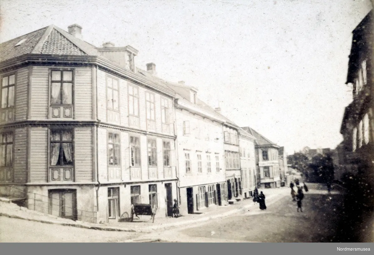 Gateparti med en hjørnegård til venstre i Storgata på Kirkelandet i Kristiansund. Ellers så ser vi mennesker i gaten lengre i bakgrunnen. Fra Nordmøre Museums fotosamlinger.