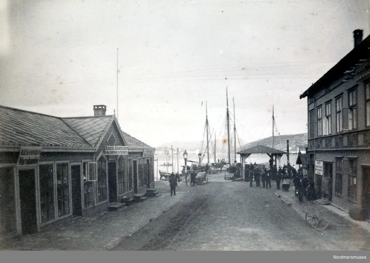 "Kirkealmenningen". Foto fra deler av bebyggelsen i Kirkeallmenningen på Kirkelandet i Kristiansund. Her ser vi blant annet Arne Arnesens Skipshandel til venstre på bildet, men Allmenningen i enden av gaten. Bildet er fra fotoalbumet "Christiansund i Fotografeir". Bildet er trolig fra 1883 etter informasjon fra Willhelm Lund samlingen. Fra Nordmøre Museums fotosamlinger.   dublett