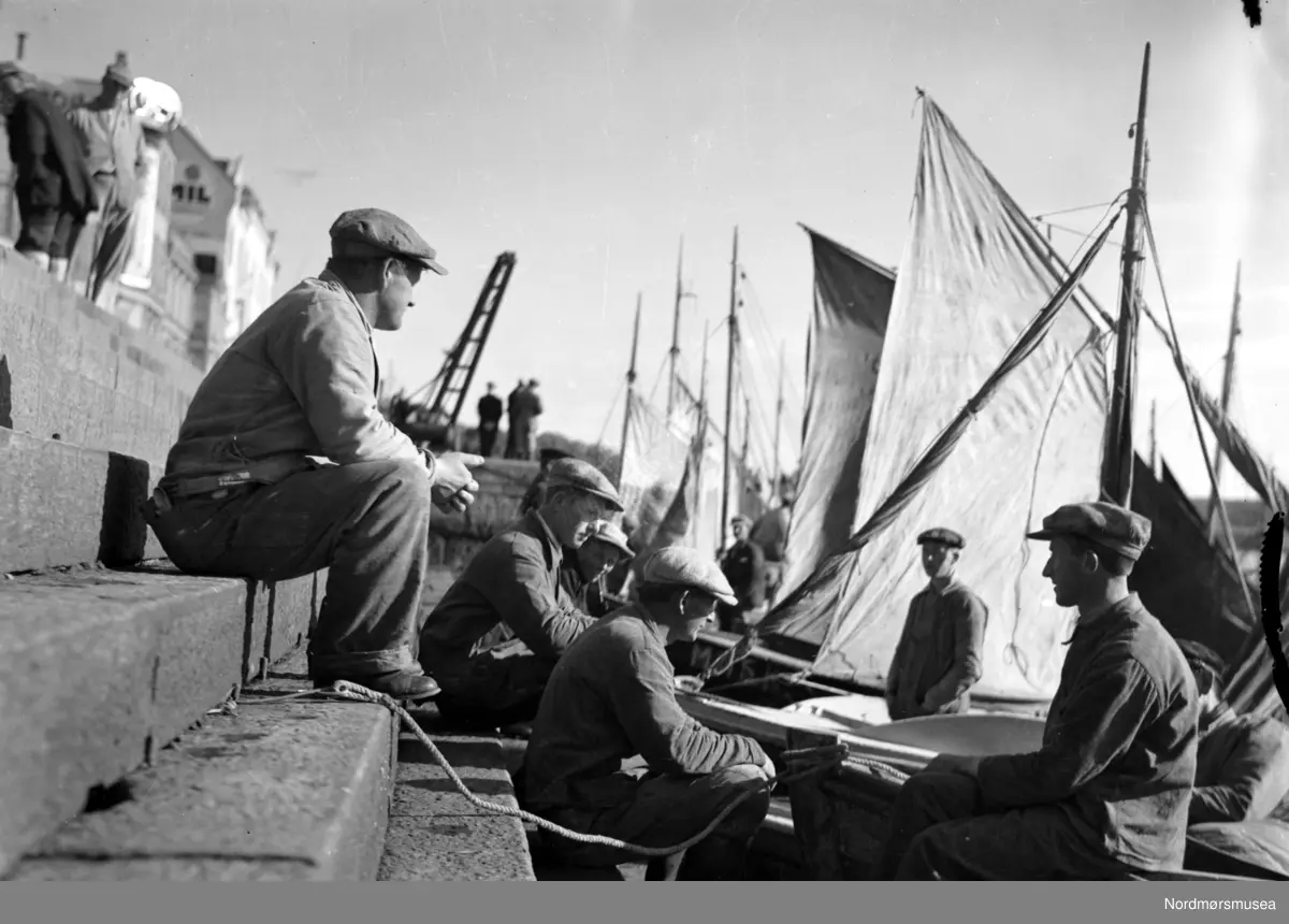 Fisktrappa i Kristiansund har vært møtested for sjøfarende og byens folk i flere generasjoner. Her fra 1930-tallet. Foto: John Myren. Film Noir-estetikk, siluett.  fiskere med sine fartøy, ved fisketrappa på Kirkelandet i Kristiansund. merk kranene langs havna. Fra Nordmøre museums fotosamlinger.
