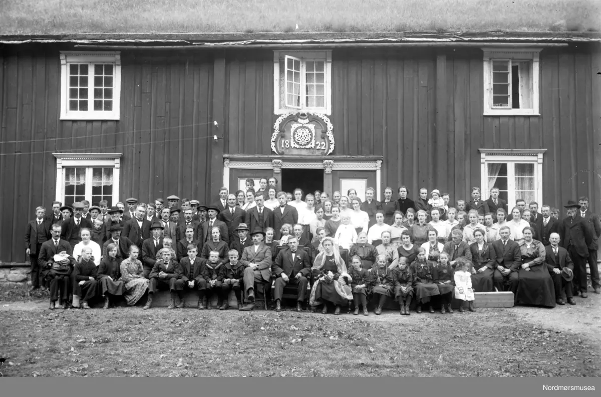 Wedding of Anne Ingebrigtsdatter (Wennevold) Hoas (1893-1976) and John Ivarsen Hoas (1891-1972), in 1921. Ingebrigt Olsen Wennevold (1858-1948), the father of the bride, Anne Ingebrigtsdatter (Wennevold) Hoås (1893-1976), is sitting at left of and next to the groom, John Ivarsen Hoås (1891-1972). I believe this is at Wennevold, June 1921. (Info: Brandon Henderson 2018) Foto fra et bryllup på Åker i Sunndal kommune. Wedding of Anne Ingebrigtsdatter (Wennevold) Hoås (1893-1976) and John Ivarsen Hoås (1891-1972), in 1921. (Info fra Brandon Henderson 2018).  Merk dørpryden med årstallet 1822 innskrevet.  Se også reg. nr. KMb-1943-000.0508 samt KMb-2007-007.0084. Fra Nordmøre Museums fotosamlinger.