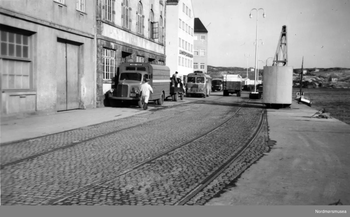 Foto fra området 
på sjøsida av det gamle Nordmøre Meieri
 på Storkaia 
ved havna på Kirkelandet i Kristiansund. 
Det er også verdt å merke seg skinnene som er lagt ned i gata (kaia). 
De kan være fra jernbaneplanene da kaia ble bygd, evt. seinere bruktr til kraner. 
Det er tydeligvis rutevogner utenfor meieriet som leverer melk i spann. Den fremste lastebilen er identifiserbar. Det står Eide Auto på den. Den var lakkert blå med hvit stripe på midten. Det er en Mercedes-Benz, sannsynligvis en 1952-modell som var registrert T-6714. Den var i trafikk til 1960. Eide Auto hade godsruter Eide-Kristiansund fra 1940 (personrute fra 1941). Ruta gikk over Gjemnes, fra 1952 over Gjemnes-Kvitnes. (Info: Sveinung Berild - 12.11.2016).  Datering av bildet er trolig omkring 1950 til 1960. Foto: Ingvard Sverdrup. Fra Nordmøre museums fotosamlinger.