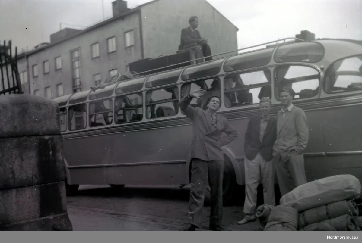 Dette kan være buss T-6708 Volvo 1952-modell med karosseri fra Bussbygg. Dette var en turbuss med 34 sitteplasser og ble mye brukt på turer i inn- og utland på 1950-tallet. (Info fra Sveinung Berild - 07.10.2016) . - Det er muligens min far, buss-sjåfør Audun Silseth som sitter på taket og antagelig venter på bagasje som skal på taket. Han kjørte mye denne bussen på 50-tallet. (Info: Finn Silseth) - Antakelig 
bussen fra Batnfjord auto vi så på bildene KMb-2008-024.N559 og KMb-2008-024.N560.  Bildet kan være fra Kristiansund. Fra Ida Knudtzons fotosamling. Russetur? bagasje, sovepose
. Fra Nordmøre museums fotosamlinger.