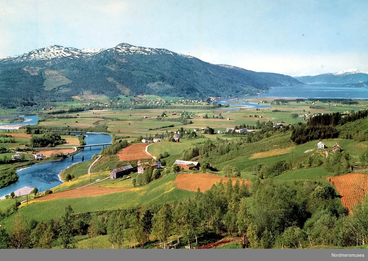 Postkort fra Surnadal, hvorfra vi ser en bygd med elv og bro. Fotograf/utgiver er Knut Aune Kunstforlag as. Fra Nordmøre museums fotosamlinger.
