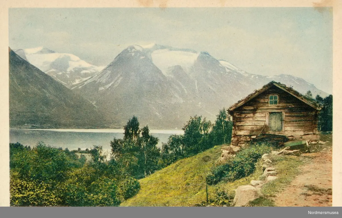 Postkort. ";Ved Strynsvandet i Nordfjord"; Foto fra Strynsvatnet, en innsjø i Stryn kommune, Sogn of Fjordane fylke, med fjellene i bakgrunnen og en gammel trebygning i forgrunnen. Datering og utgiver av postkortet er ukjent. Fra Nordmøre museums fotosamlinger.
 Reg: EFR





