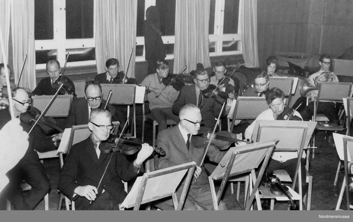 Fioliner og horn på prøve i Tempohuset 1969: Fra venstre: Anders Bergem (rutete skjorte), Bjarne Sakshaug (biller), Leif Ulvund, Johan Kjelle, Magnar Ersvik (foran), Anders Gjendem (bak), Einar Westre, Rudolf Ziegler Unhjem, Birger Andresen, NN, Ole Dahl Rossbach, Kåre Karlsen (foran), Lisa Odden og Ivar Olsen. Foto: Trygve Rossbach

Bilde fra Trygve Rossbachs dokumentasjon av Kristiansund Symfoniorkesters virksomhet på andre halvdel av 1900-tallet; ca 1950-2000. Prøver, turer, sosialt samvær og konserter. (Fra Normøre museum sin fotosamling.)
