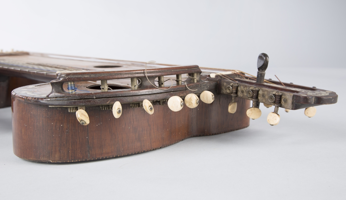 Trippelgitar, 3 halser. Lokk av gran, består av 3 deler, bunn og sarg muligens en sort mahogny. Den lengste halsen fortsetter øverst ut i en ny resonanskasse, hvis overside tjener som skruekasse for den mellomste halsen. Her er også et lydhull. En ekstra resonanseffekt oppstår ved at strengene som løper over denne resonanskassedelen under spillet svinger med. Instrumentet er utsmykket med rike perlemosrsforsiringer, delvis stekt fargede. Ornamentene er dels svært enkle og dels meget delikate blomstermønstre. Hovedlydhullet er omgitt av en 4 cm. bred krans av geometrisk intarsia, som kan minne om maurisk stil. Instrumentet har i alt 4 lydhull og 3 adskilte strengestoler. Den lengste halsen har 7 frittsvingende bass-strenger de to andre hver 6 strenger og 13 tverrbånd av metall. Runde skruehoder av elfenben. Likeså oversadler og strengestoler av elfenben.
Usignert.