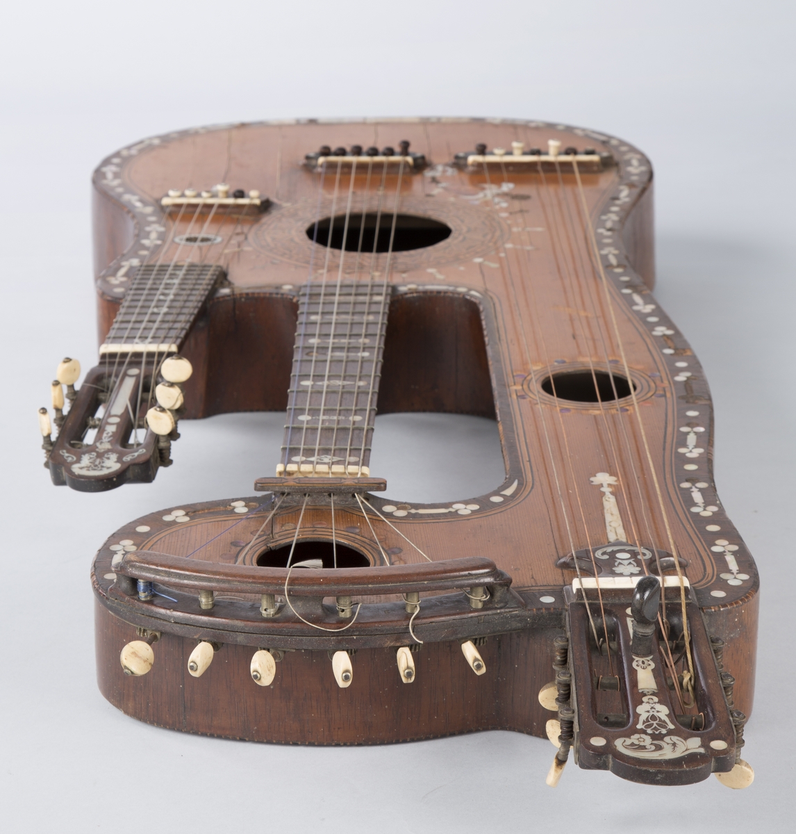 Trippelgitar, 3 halser. Lokk av gran, består av 3 deler, bunn og sarg muligens en sort mahogny. Den lengste halsen fortsetter øverst ut i en ny resonanskasse, hvis overside tjener som skruekasse for den mellomste halsen. Her er også et lydhull. En ekstra resonanseffekt oppstår ved at strengene som løper over denne resonanskassedelen under spillet svinger med. Instrumentet er utsmykket med rike perlemosrsforsiringer, delvis stekt fargede. Ornamentene er dels svært enkle og dels meget delikate blomstermønstre. Hovedlydhullet er omgitt av en 4 cm. bred krans av geometrisk intarsia, som kan minne om maurisk stil. Instrumentet har i alt 4 lydhull og 3 adskilte strengestoler. Den lengste halsen har 7 frittsvingende bass-strenger de to andre hver 6 strenger og 13 tverrbånd av metall. Runde skruehoder av elfenben. Likeså oversadler og strengestoler av elfenben.
Usignert.