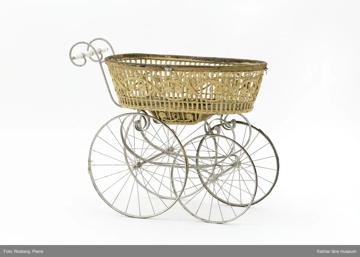 KLM 28338:1. Barnvagn, liggvagn. Hjul och underrede av metall målade i silverfärg, troligen ej ursprunglig. Två hjul är gummiklädda, ett saknar detta och det fjärde är försett med flätat naturmaterial. Handtag av vitt porslin. Vagnskorg med trästomme bestående av flätade stående och liggande spjälor målade i ljusgul nyans. Övre kanten av korgen är klädd med grovt ylletyg och på utsidan ett vävt tygband i gul ylle. Korgens botten är klädd med grönt konstläder och säckväv målat i rutmönster. I mitten av korgen en försänkning. På korgens sidor fästen för sufflett. På hjulen instansat: BRENNABOR D.R.G.M.