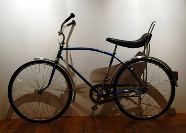 Pojkcykel med "limpa-sadel", ca 1970-tal. Märke: Maraton AB J Benson Eskilstuna Malmö. Blå ram, gul och blå dekor samt svart och vit text "MARATON" på ramen. Högstyre, lång svart "limpa-sadel", cykelställ, blocklås, bakljus, reflexer på tramporna samt aluminiumskärmar. Däcken är av grövre, dubbad typ.