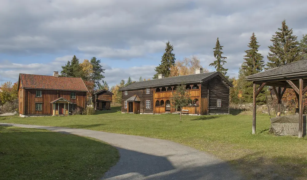 Grusveg som fører over et tun med en låve lengst til høyre, samt to brunlige tømmerbygninger hvorav det ene med svalgang.