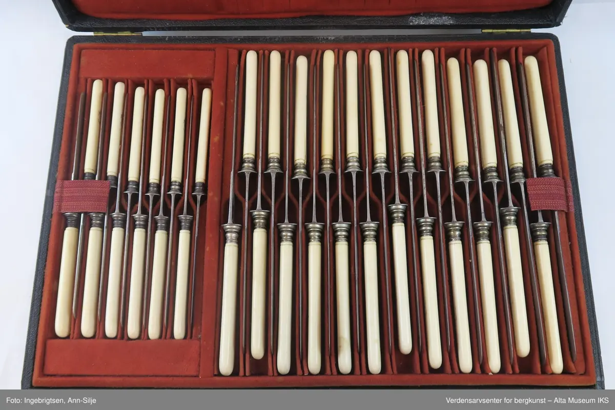 Rektangulær koffert med 12 mindre kniver (til frokost) og 24 større kniver (til middag). Knivene ligger pent anrettet i rødtrukket bestikkasse. Knivene har hvite håndtak og metalfarget knivblad.