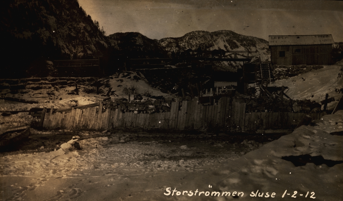 Storstrømmen sluse. Nedre Fangdam. Vinter, snø, is og en elv. Fjell i bakgrunn. Bro med hester og menn på. En bygning/hus i høyre billedkant. (Arkiv: Kanalvesenet/Vassdragsvesenet Uba-2-05)