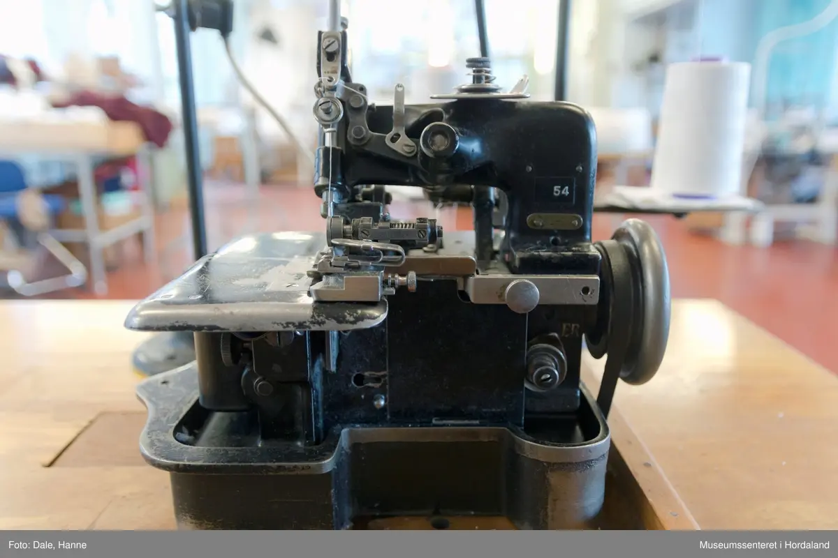 Ei to-tråds Singer symaskin til falding/kantar på strikka stoff, med original motor, lampe, montert på bord. 

Modell 81-6, merka med nummer 54. Maskinnummer AB503010. Overlockmaskin med faldeapparat, til kanting av stoff.