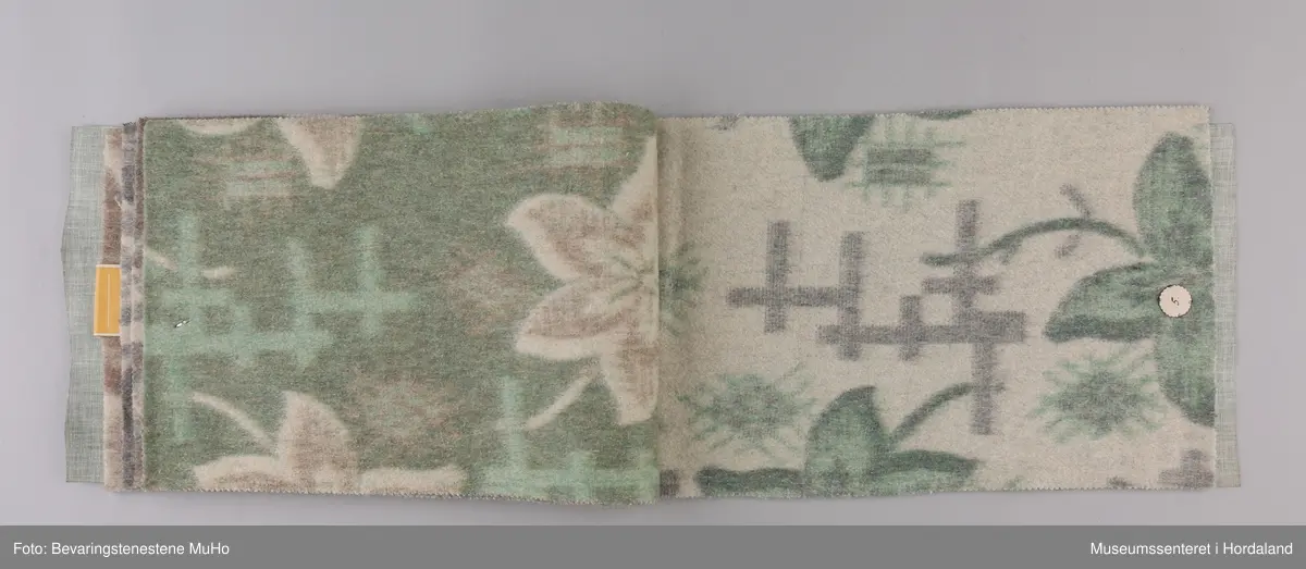 Stoffprøvebok frå Salhus Væverier. Prøver på ullteppe med blomstermotiv, feste i papp/hyssing. Omslag i grønt plastbelagt stoff i skinnimitasjon/skai.