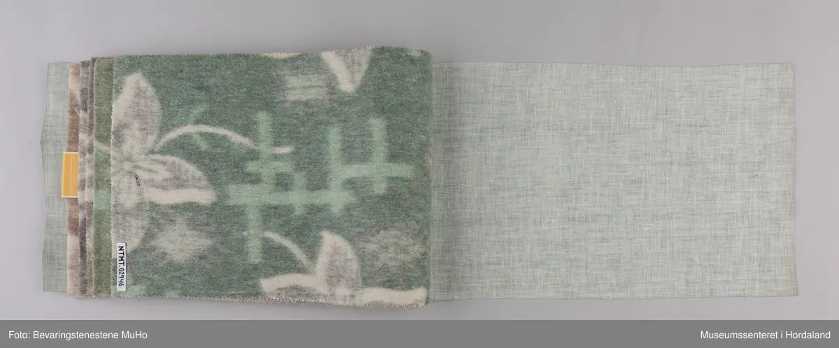 Stoffprøvebok frå Salhus Væverier. Prøver på ullteppe med blomstermotiv, feste i papp/hyssing. Omslag i grønt plastbelagt stoff i skinnimitasjon/skai.