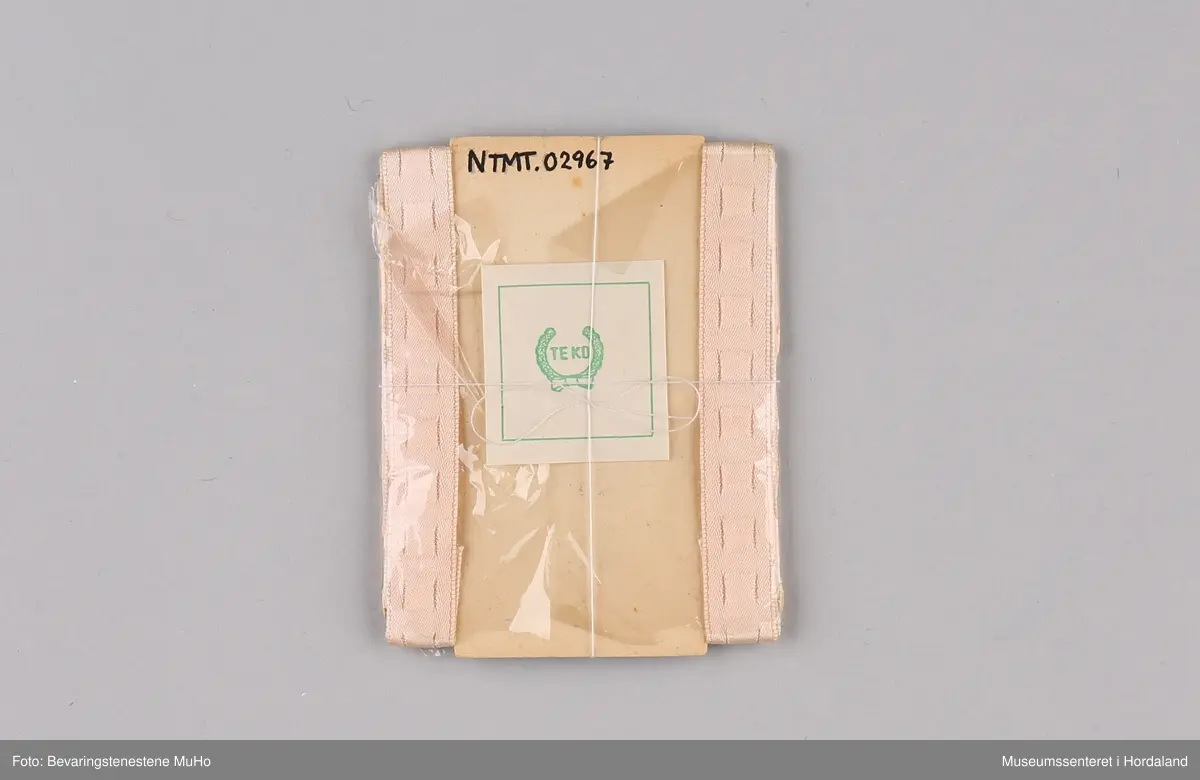 Ferskenfarga "Tyx" skulderstropper til BH, i tekstil og elastikk med metallhekter. Norskprodusert av Teko fabrikker i Bergen, pakka i papp- og plastemballasje.