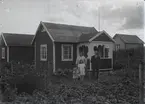 En stuga, man, kvinna och barn i Kalmar södra koloniområde, fotograferat omkring 1930. Kalmar södra koloniförening grundades 1917 och har idag 105 kolonilotter. Området ligger strax söder om länssjukhuset i Kalmar med huvudingång från Stensbergsvägen.