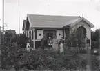 En stuga, man, kvinna och barn i Kalmar södra koloniområde, fotograferat omkring 1930. Kalmar södra koloniförening grundades 1917 och har idag 105 kolonilotter. Området ligger strax söder om länssjukhuset i Kalmar med huvudingång från Stensbergsvägen.