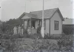 En stuga, två män och två kvinnor i Kalmar södra koloniområde, fotograferat omkring 1930. Kalmar södra koloniförening grundades 1917 och har idag 105 kolonilotter. Området ligger strax söder om länssjukhuset i Kalmar med huvudingång från Stensbergsvägen.