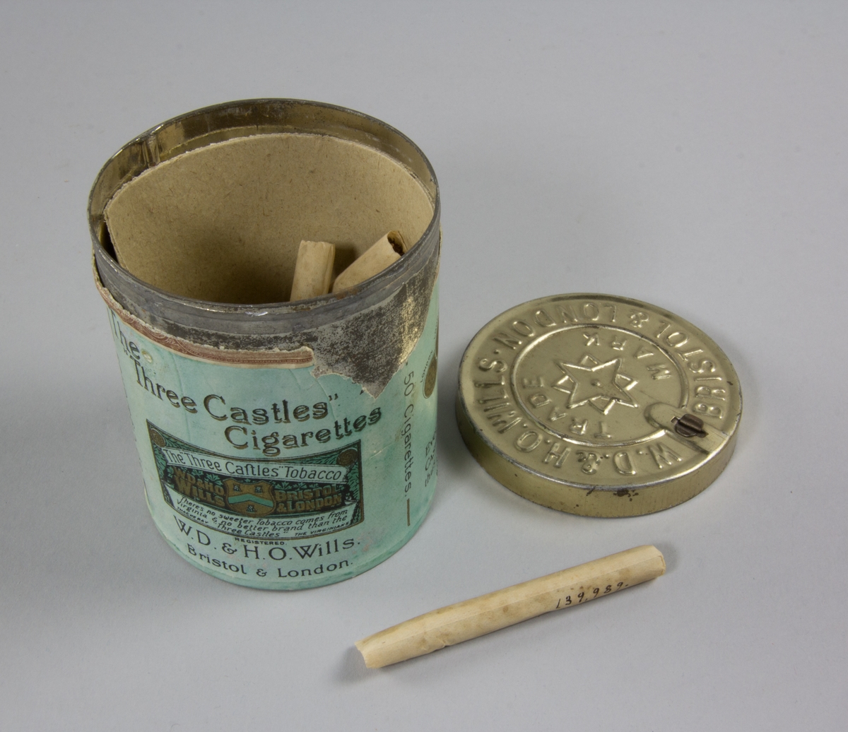 Burk, cylindrisk, av plåt med plant avtagbart lock. Präglad text på locket. Runt burken påklistrad pappersetikett. Burkens insida pappfodrad. Burken innehåller fem cigaretter.