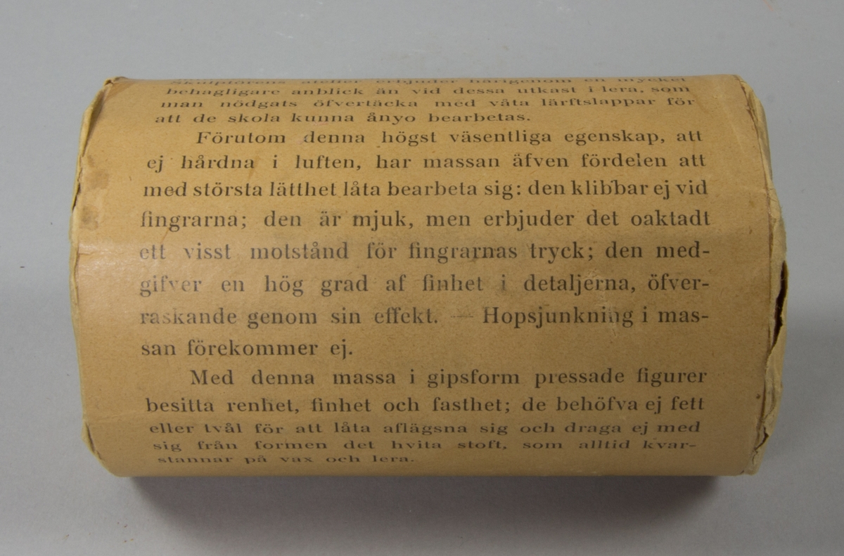 Plastelinamaterial, 6 st. 4 st cylindriska i obrutna förpackningar med brunt omslagspapper och lång reklamtext. 2 st utan omslagspapper och använda till häften. Ovanpå en av dem klumpar av plastelina.