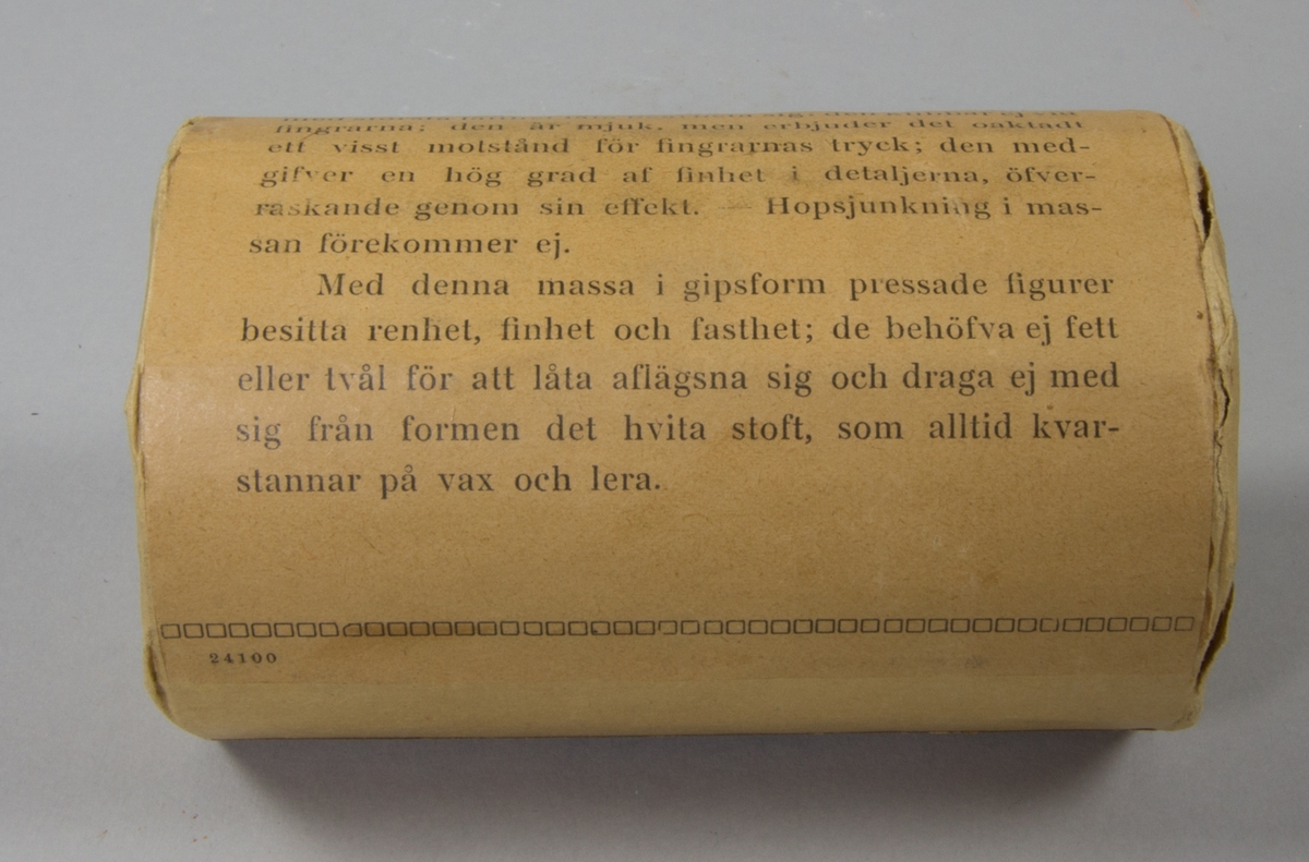 Plastelinamaterial, 6 st. 4 st cylindriska i obrutna förpackningar med brunt omslagspapper och lång reklamtext. 2 st utan omslagspapper och använda till häften. Ovanpå en av dem klumpar av plastelina.