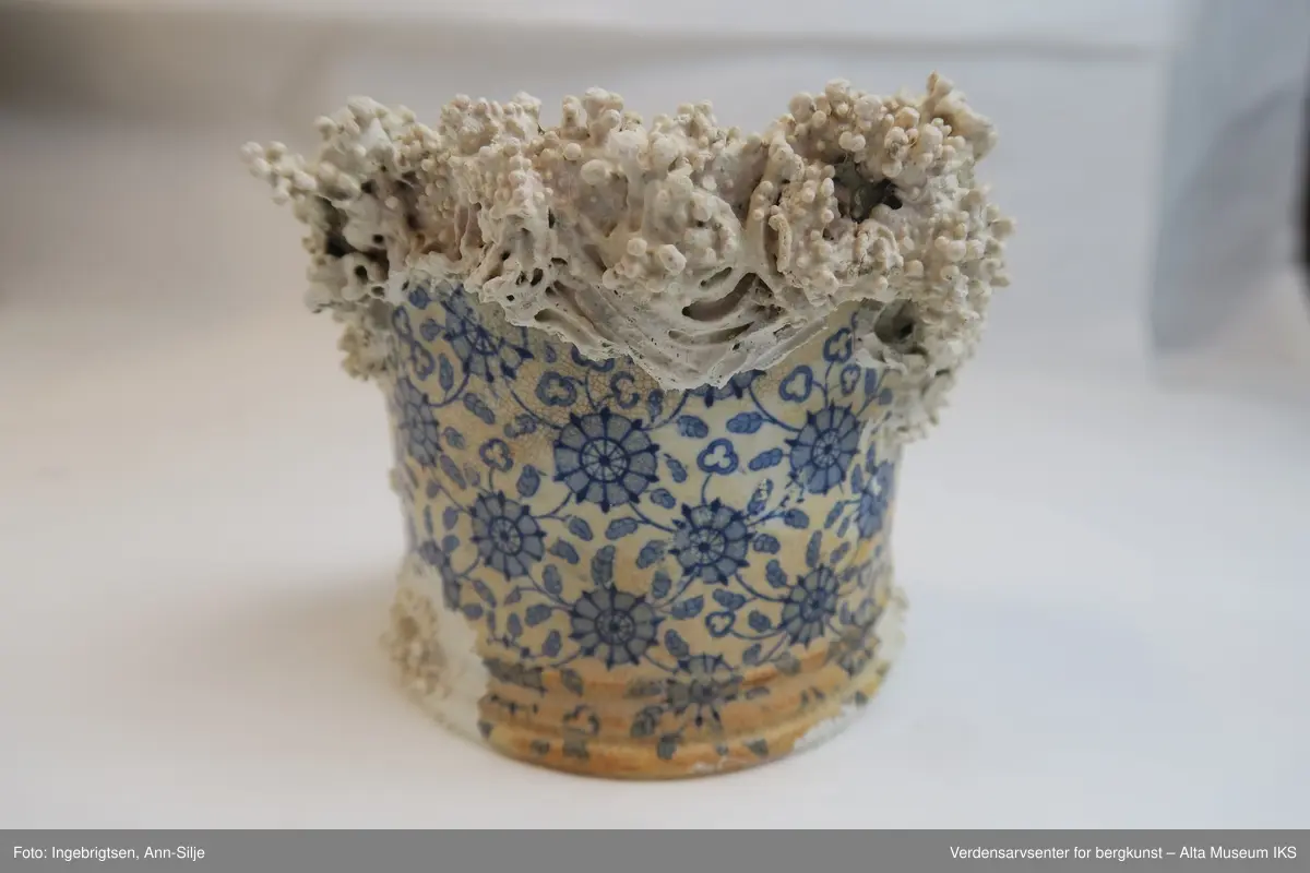 Sylinderformet beholder, krus, som har blitt knust. Gjenstanden er dekorert med blomstermotiv - både utenpå og inni. Dekket delvis av rur.