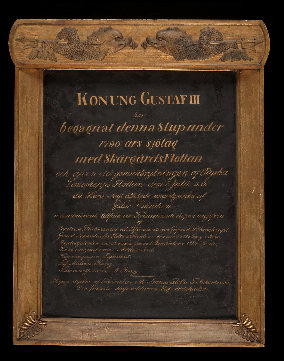 Minnestavla i koppar, svartmålad med inskription i guldskrift. Ram av förgyllt trä. Överstycke med 2 delfiner förande neptunigaffel.