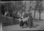 Här är Elna Brundin ute på en vårlig promenad med Sören i vagnen och Eva framför. Bilden tagen ca 1923, på våren. Huset i bakgrunden är troligen västra gaveln på Grand Hotel och till vänster ovanför staketet ligger Frimurarhuset.