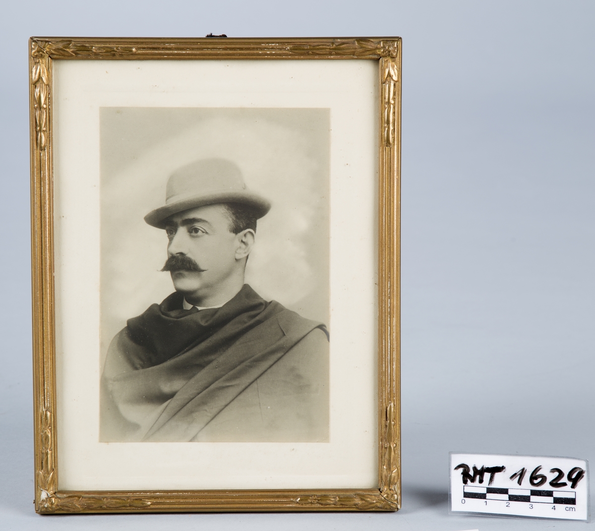 Svart-hvitt fotografi satt i glass og ramme. Rammen har relieffmønster og er forgylt.